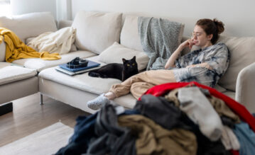 Junge Frau liegt mit ihrer Katze auf der Couch und neben ihr liegt ein großer Haufen Wäsche zum Zusammenlegen