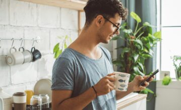 Ein Mann blickt mit seiner Kaffeetasse in der Hand auf sein Handy.