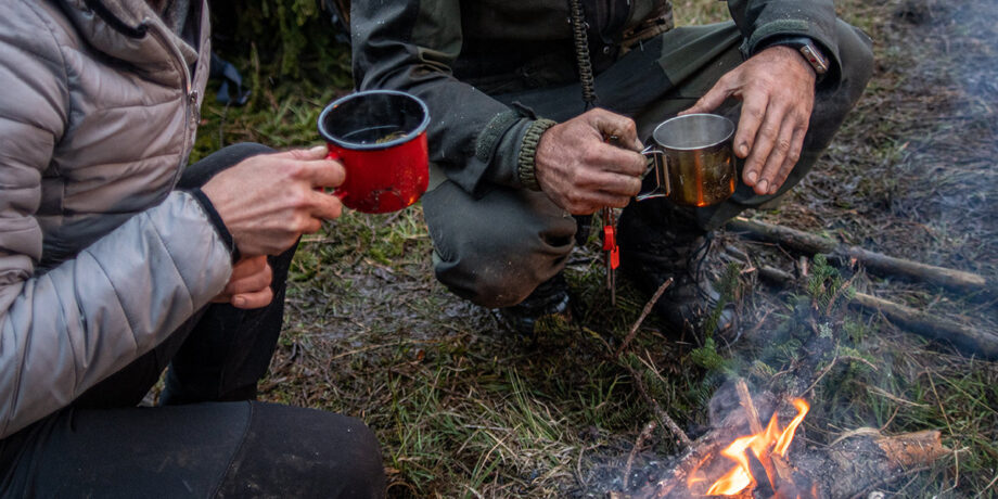 Zwei Abenteurer hocken vor einem Lagerfeuer und trinken ihren Kaffee