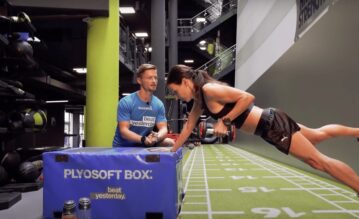 Eine Frau macht unter Anleitung eines Trainers ein Krafttraining im Fitnessstudio. Sie stützt sich auf eine Plyosoft Box und führt eine Ruderübung mit Kurzhanteln aus.