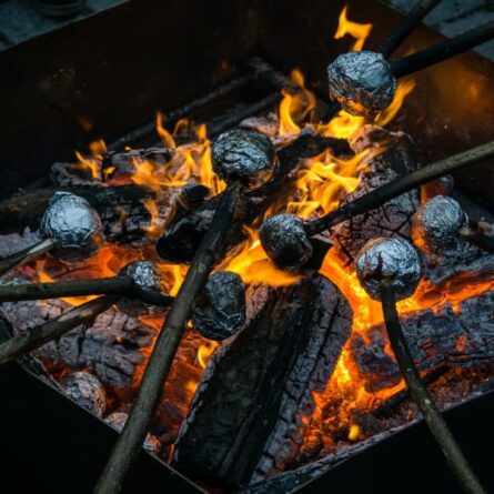 Stöcker mit Champignons, die in Alusfolie gewickelt sind, werden über ein Feuer gehalten