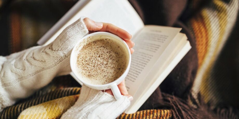 Frau mit Strickpulli hat ein Buch auf dem Schoß und eine Tasse Kaffee in der Hand und entspannt zuhause