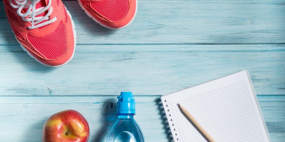 Laufschuhe, ein Apfel, eine Wasserflasche und Zettel und Stift liegen auf einem Holztisch