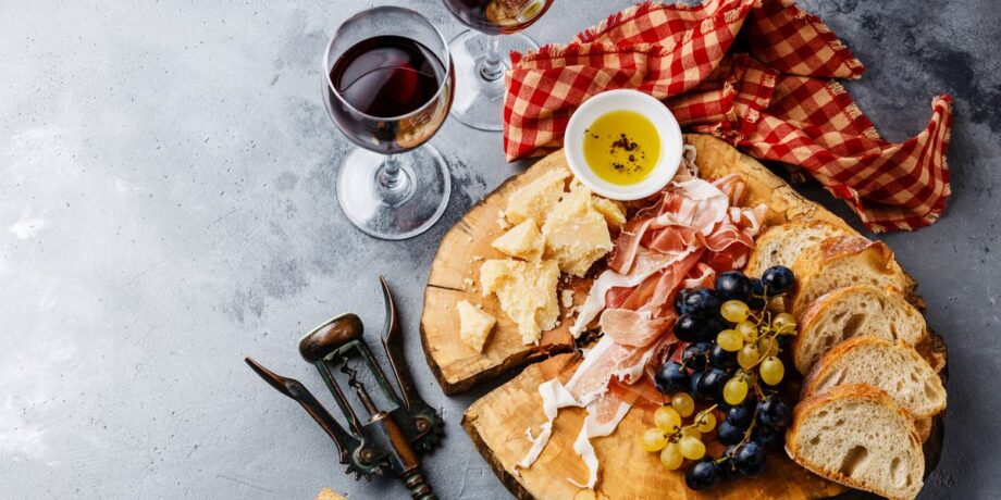 Holzteller mit Brot Käse, Weintrauben, Schinken und daneben ein Glas mit Rotwein gefüllt