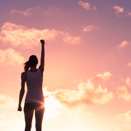 Frau steht vor einem Sonnenaufgang und streckt motiviert ihren Arm in die Luft