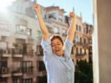 Junge Frau steht morgens auf ihrem Balkon in der Sonne und stretcht sich