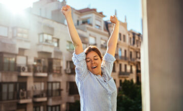 Junge Frau steht morgens auf ihrem Balkon in der Sonne und stretcht sich