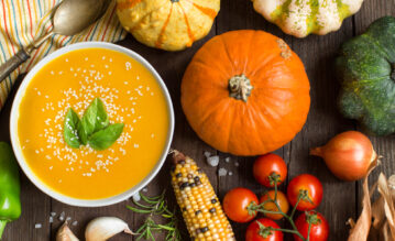 Eine Schale mit Kürbissuppe, unterschiedliche Kürbissorten und weiteres Herbstgemüse angerichtet auf einem Holztisch.