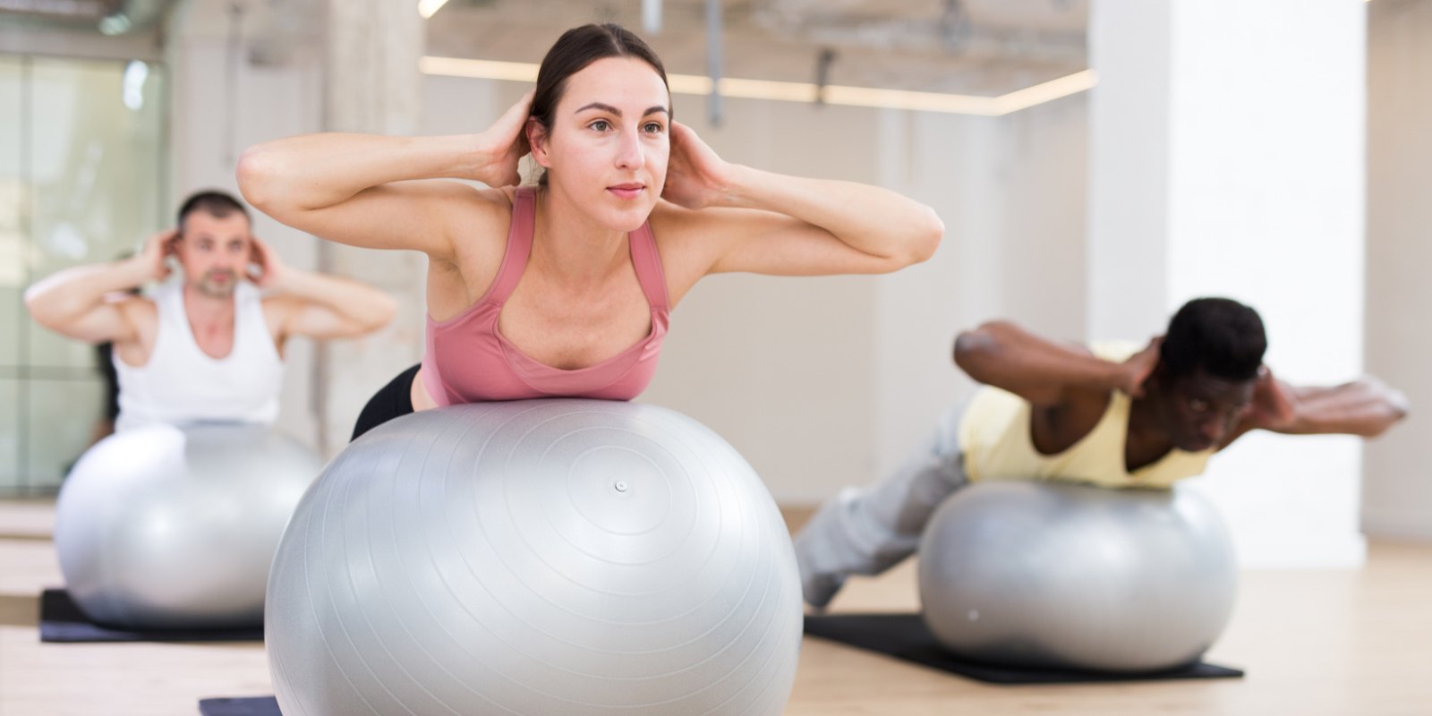 Junge Frau im Sportoutfit liegt mit dem Bauch auf einem Gymnastikball, hat ihre Hände am Hinterkopf und zieht ihren Oberkörper nach oben, um eine gerade Linie zu bilden.