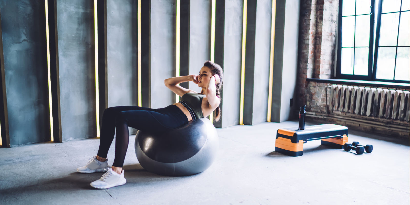 Junge Frau im Sportoutfit liegt mit dem Rücken auf einem Gymnastikball und macht Sit Ups.