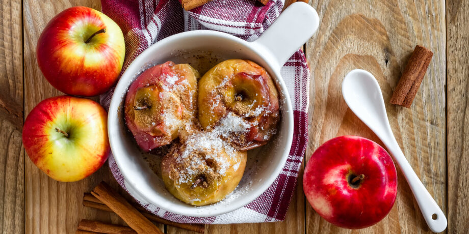 Gebackene Äpfel mit Zimt und Zucker bestreut in einer weißen Schüssel, neben frischen Äpfeln und Zimtstangen auf einem Holztisch.