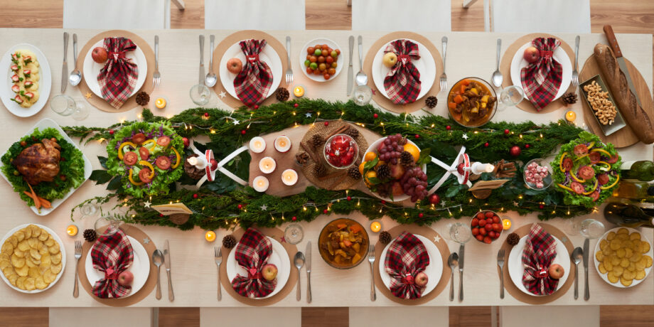 Weihnachtlich dekorierter Esstisch von oben betrachtet mit Kerzen, Tannenzweigen und verschiedenen Gerichten wie Braten und Gemüse.