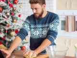 Ein Mann mit einem festlichen Pullover steht in einer Küche und knetet Teig auf einer Arbeitsplatte, im Hintergrund ist ein geschmückter Weihnachtsbaum zu sehen.