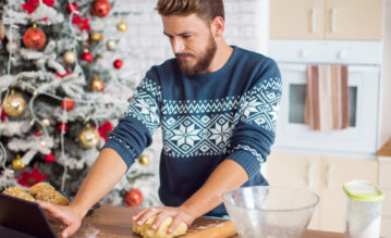 Ein Mann mit einem festlichen Pullover steht in einer Küche und knetet Teig auf einer Arbeitsplatte, im Hintergrund ist ein geschmückter Weihnachtsbaum zu sehen.