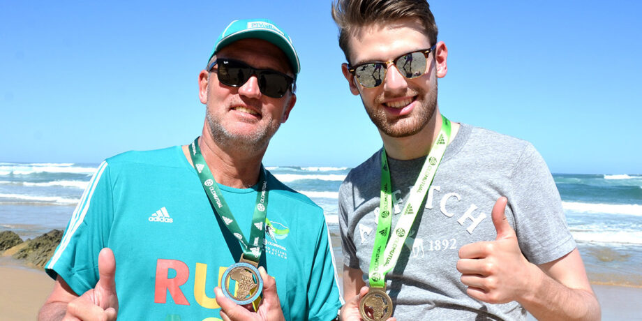 Friedhelm Weidemann steht mit seinem Sohn am Strand und hält Medaillen in die Kamera