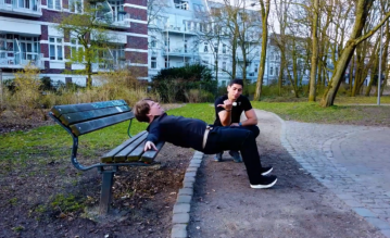 Faul wird fit Teilnehmer Colin macht Fitnessübungen an einer Parkbank während Trainer Gino daneben hockt und Tipps gibt