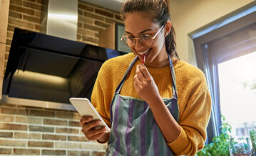 Junge Frau steht in der Küche und schaut fröhlich auf ihr Handy, um ein Rezept zu lesen.