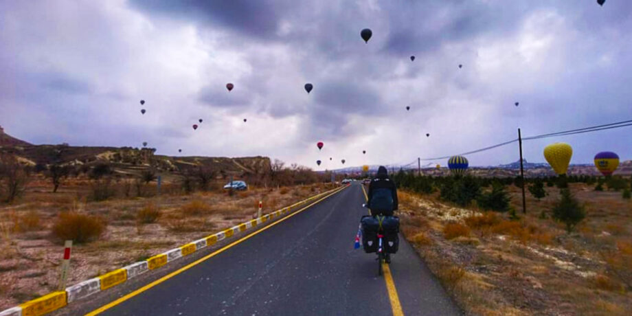 Anika Brust und Denis Scharnow fahren mit dem Fahrrad eine Straße entlang. In der Ferne sieht man hunderte Heißluftballons am Himmel.
