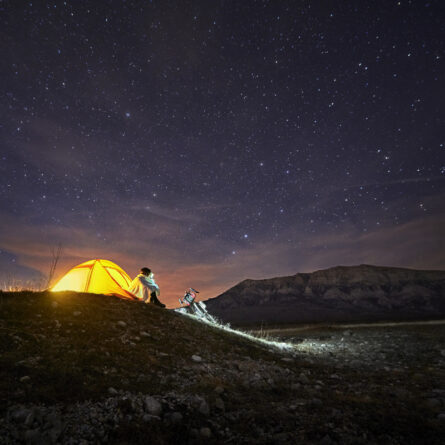 Mann sitzt nachts vor seinem beleuchteten Zelt auf einem Berg