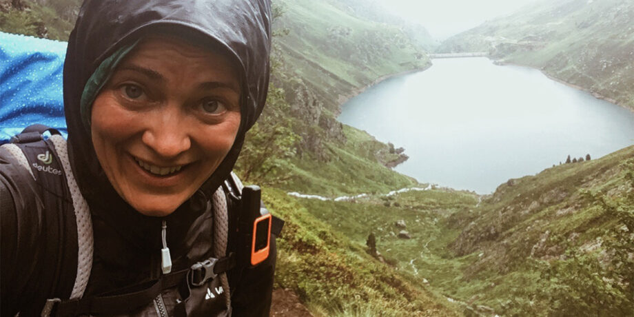 Ana Zirner lächelt durchnässt bei einer Wanderung in die Kamera. Im Hintergrund liegt ein Bergsee.