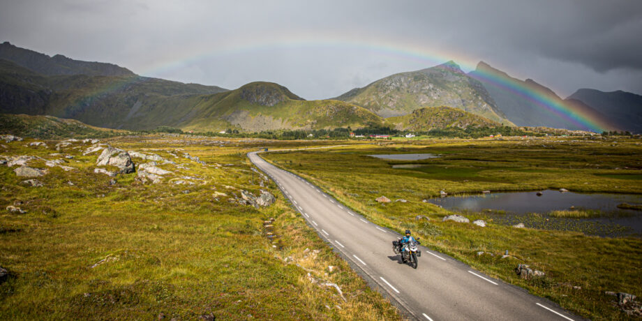 Maia fährt mit ihrem Motorrad durch Norwegen und hinter ihr ist ein Regenbogen am Himmel zu sehen