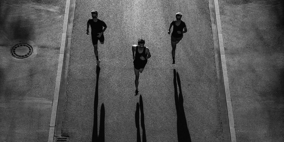 Schwarz-weiß-Foto mit 3 Läufern, die große Schatten auf die Asphaltstraße werfen
