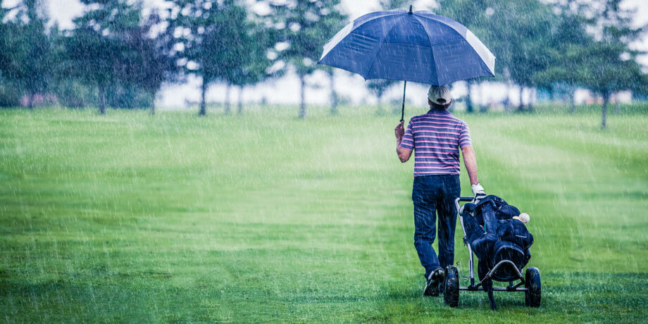 Golfer zieht seinen Trolly bei Regen über den Platz und hält einen Regenschirm in der Hand