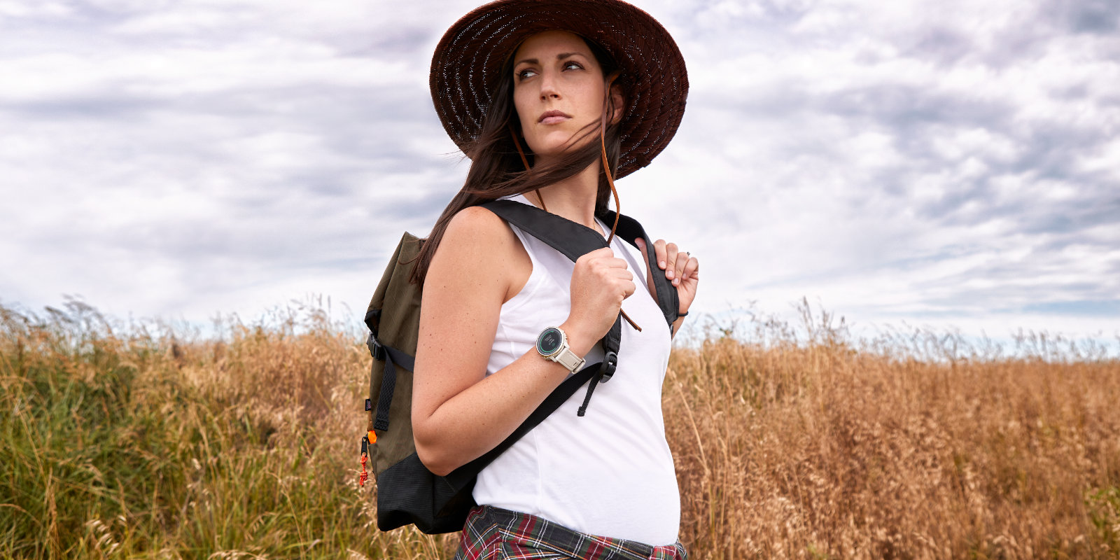 Frau mit Hut und Garmin Uhr am Handgelenk wandert Wochen nach der Geburt durch ein Feld