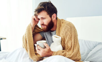 Mann mit Leisure Sickness Syndrom sitzt krank im Bett mit Tee und Wärmflasche