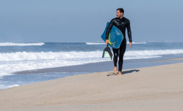 Mann geht mit seinem Bodyboard und Flossen am Strand entlang und guckt auf das Meer