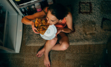Frau hat Stress und sitzt spät abends vor dem Kühlschrank und isst Pizza