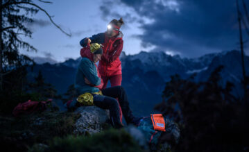 Eine Frau in roter Jacke legt einer verletzten Person in gelber Jacke eine Rettungsdecke an, nachts auf einem Felsvorsprung mit Bergen im Hintergrund.