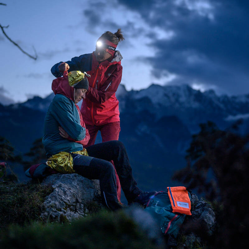 Eine Frau in roter Jacke legt einer verletzten Person in gelber Jacke eine Rettungsdecke an, nachts auf einem Felsvorsprung mit Bergen im Hintergrund.