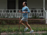 Ein Mann joggt auf einem Weg vor einem weißen Haus mit Veranda. Er trägt ein blaues Langarmshirt, graue Shorts und weiße Laufschuhe. An seinem Handgelenk trägt er eine Fitnessuhr. Im Vordergrund sind ein niedriger Zaun und grüne Pflanzen zu sehen.