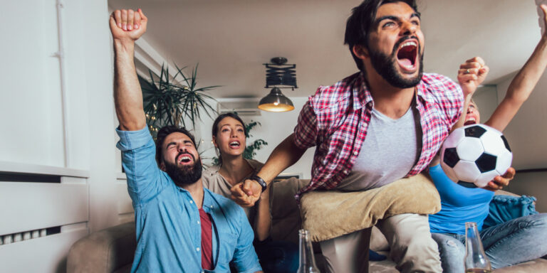 Männer und Frauen sehen sich zuhause im Wohnzimmer ein Fussballspiel an und jubeln vor Freude