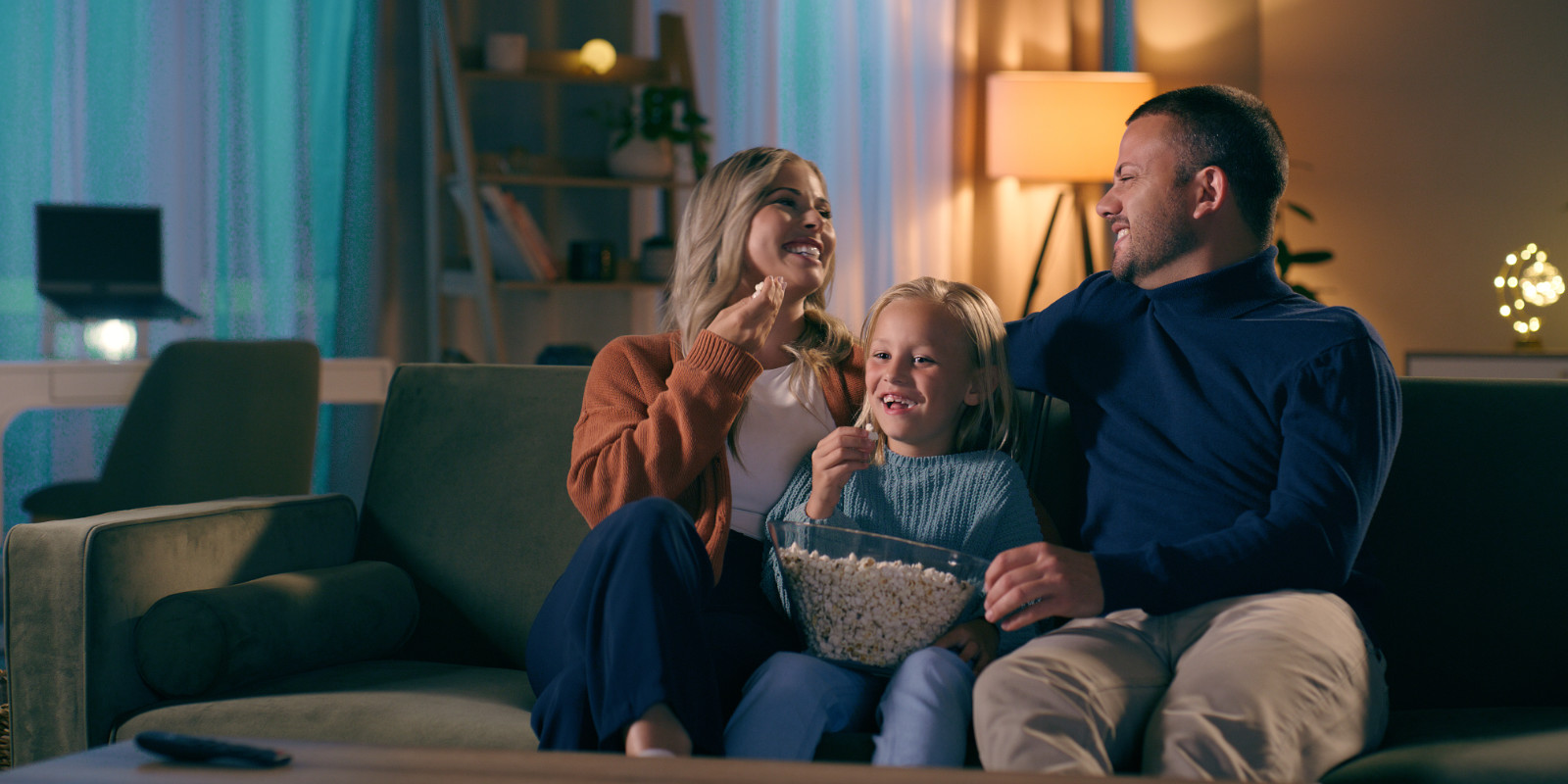 Familie sitzt gemeinsam mit ihrem Kind auf der Couch bei einem Filmabend und isst Popcorn