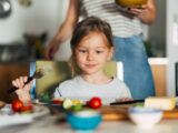 Kleines Mädchen sitzt mit ihren Eltern am Frühstückstisch und schaut erfreut auf das Essen