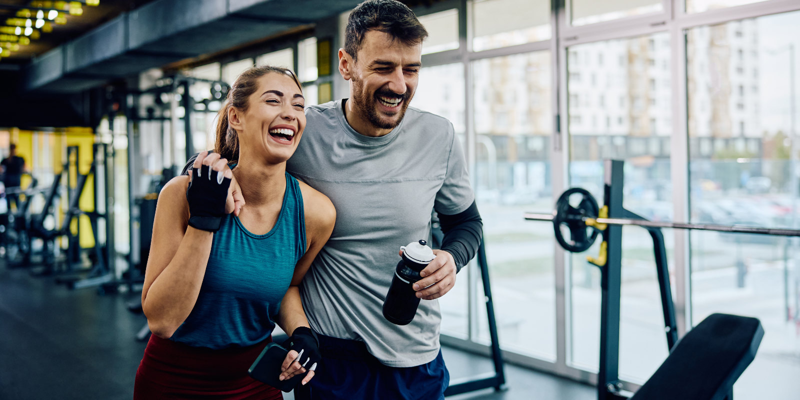 Mann hat den Arm um eine Frau gelegt und geht mit ihr lachend durch das Fitnessstudio