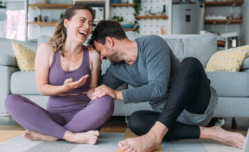 Frau und Mann sitzen auf dem Boden im Wohnzimmer in Sportbekleidung und lachen
