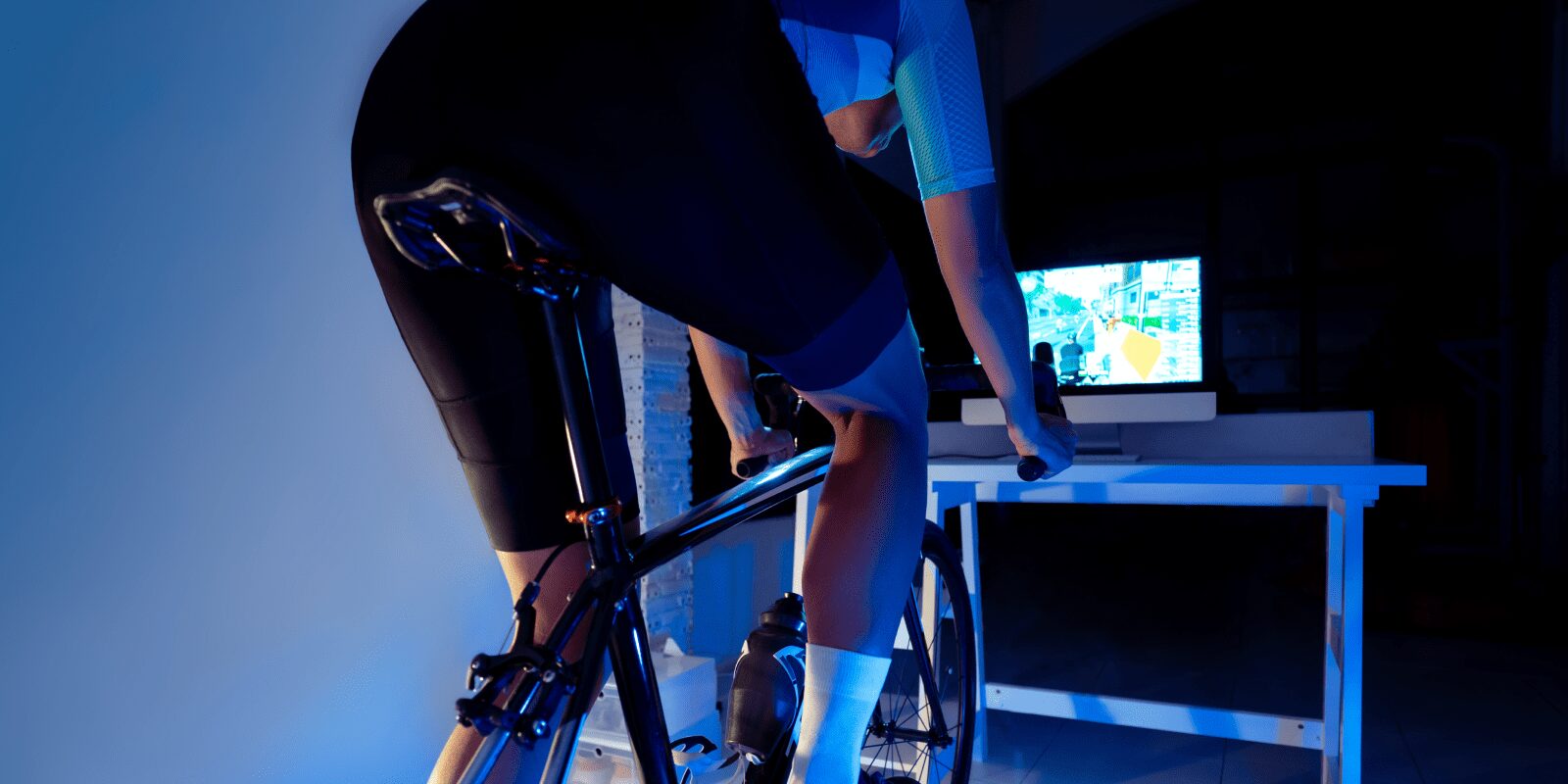 Eine Person sitzt in Sportkleidung auf einem Rollentrainer und blickt auf eine virtuelle Radstrecke vor sich auf einem Laptop.