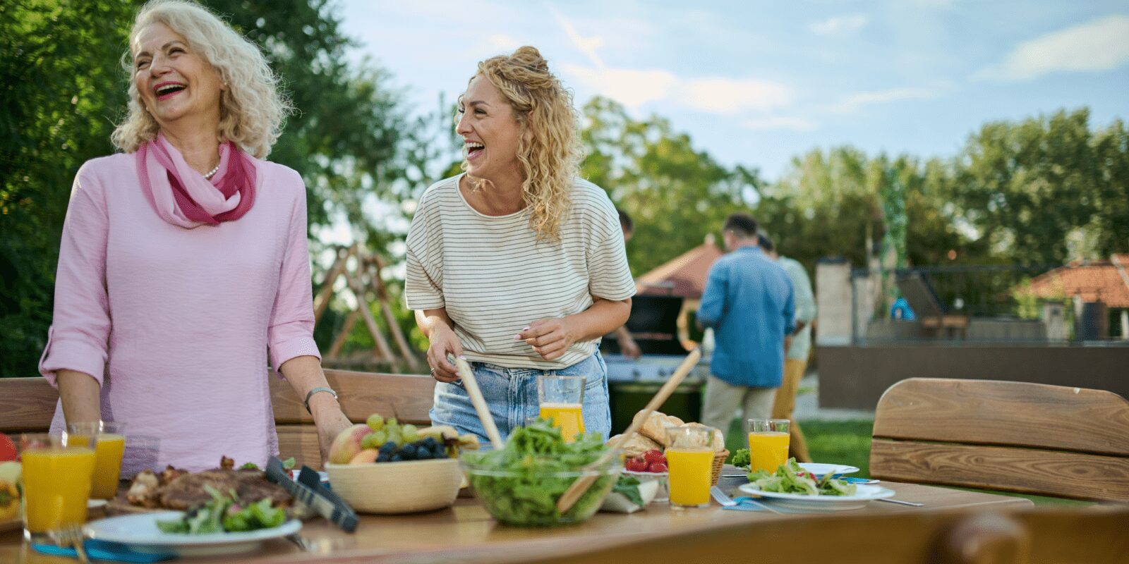 Zwei Frauen bereiten lachend Essen für einen Grillabend vor. Im Hintergrund stehen Männer am Grill.