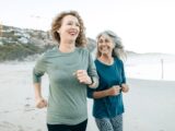 Zwei ältere Frauen laufen lachend am Strand entlang.