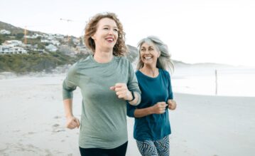 Zwei ältere Frauen laufen lachend am Strand entlang.