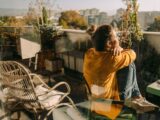 Eine Frau sitzt auf ihrem Balkon in der Sonne und schaut in die Ferne.