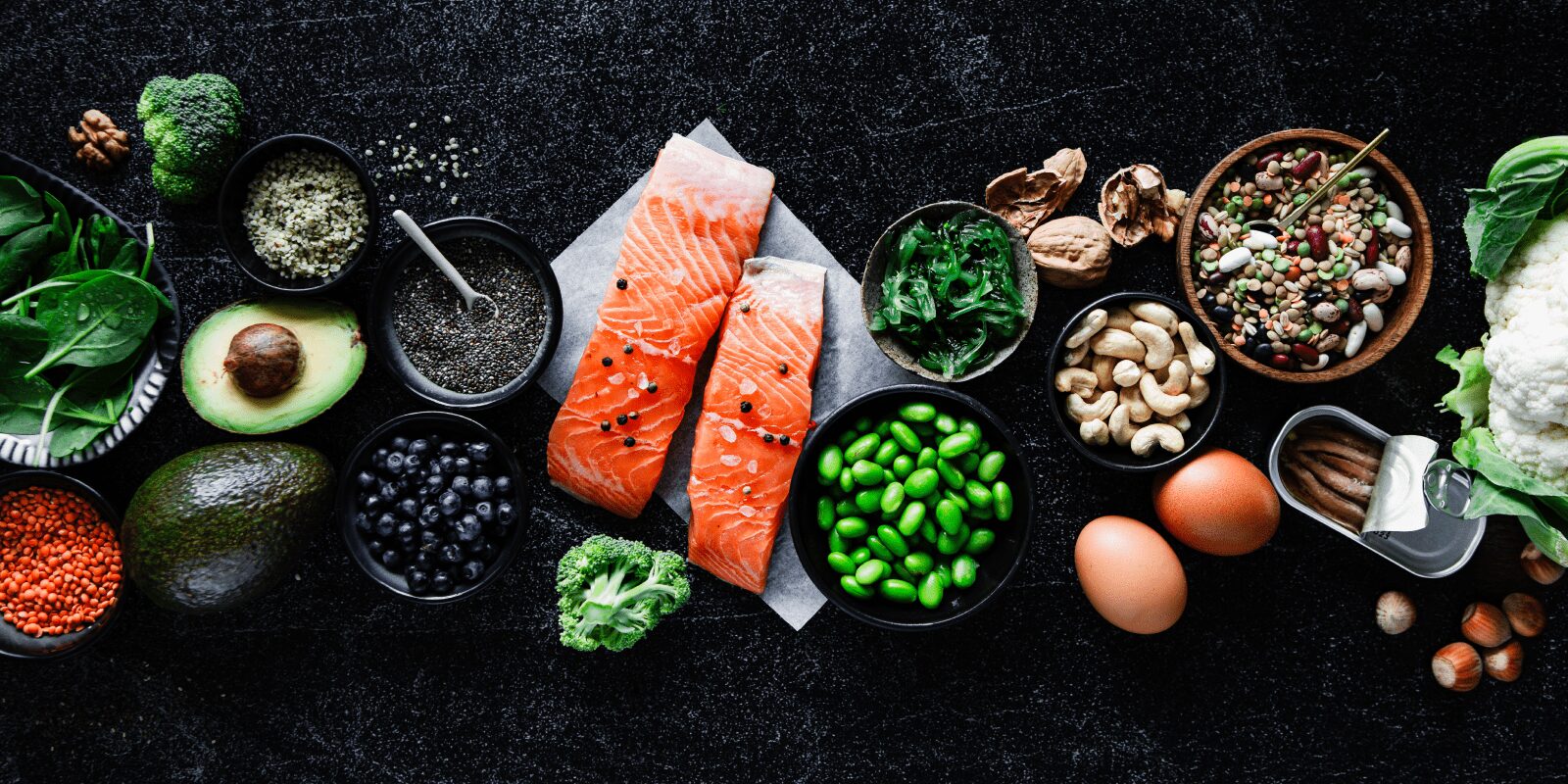 Lebensmittel wie Fisch, Bohnen, Nüsse oder Eier liegen nebeneinander auf schwarzem Untergrund.