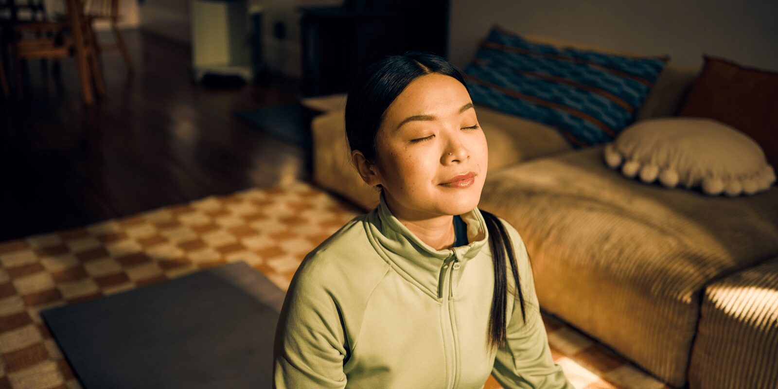 Eine junge Frau sitzt im Sonnenlicht auf ihrer Yoga-Matte und hat die Augen geschlossen.