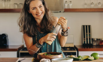 Reife Frau steht fröhlich in der Küche und würzt ihr Essen